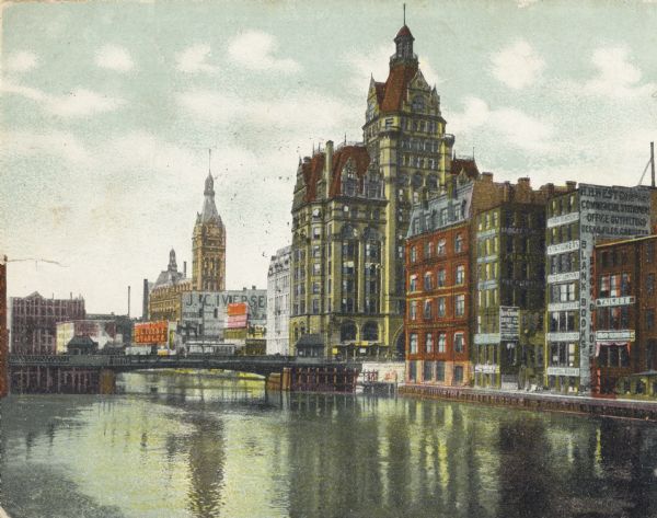 View down river toward a bridge. On the right are buildings lining the river with billboards and signs.
