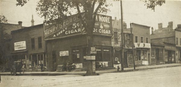 Northeast corner of the intersection, future site of the Stephenson Building.  A cigar store stands on the corner, with a harness store on the left, and more storefronts on the right.  A tree with attached signs is on the corner next to the sidewalk.  A carriage is parked in front of the harness shop.