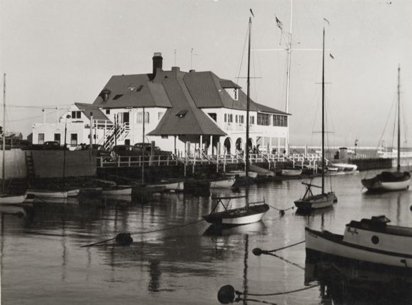 Exterior of building from shoreline looking out to Lake Michigan. Along the pier are numerous docked boats and sailboats moored in water. On the left automobiles are parked along a fence.