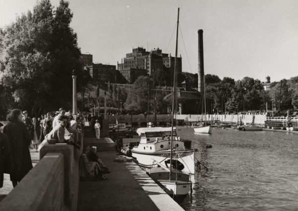 Basin or slip area.  The city is on a hill in the background, and boats are in the river.  On the left is a sidewalk and railing with stairs. People are walking or sitting at various points along the railing.