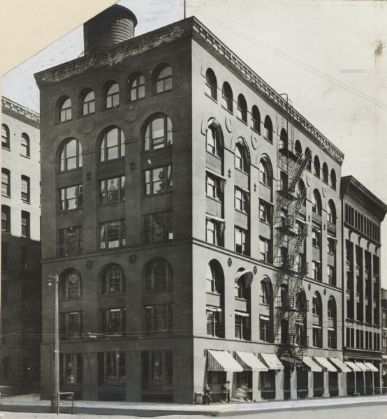 Former home of the <i>Milwaukee Journal</i> newspaper, at the corner of Michigan and Milwaukee Streets. A fire escape goes up the middle of the building on the right, and a water tower drum is on the roof. Awnings are over the first floor windows on the right, and two men are on the sidewalk near the corner.