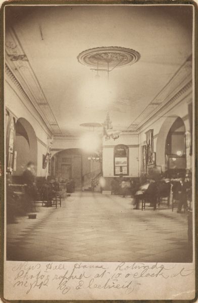 Interior view of lobby area with several people sitting in chairs.  Manuscript notations on the mount makes much of the fact that it was photographed at night by electric light and adds ambiguously "the first electric illumination" (in Milwaukee?).