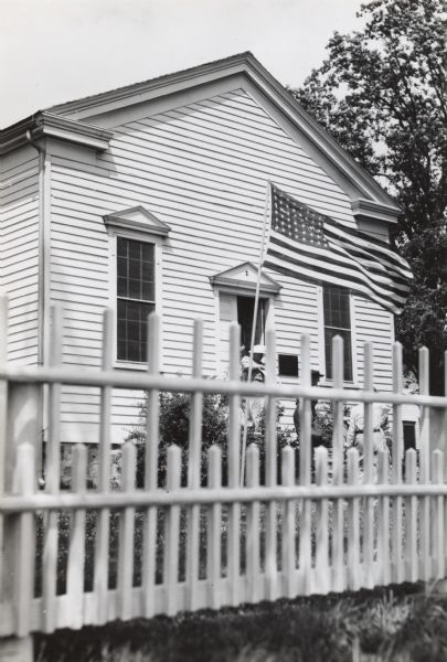 A flag is flying on a flagpole in the front yard near the entrance of the Greek Revival building. Several people are exiting the one story building. A white fence is in the foreground.