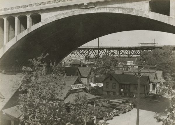Small neighborhood of the city at the west end of the Menomonee River Valley.  Overhead is the second Wisconsin Avenue Viaduct, demolished in 1992.  Cars are parked outside the Seven Eleven tavern, and in the background a trolley goes by on an elevated track.