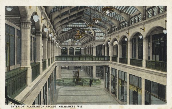Interior of the arcade from the second floor. There are skylights and chandeliers in the ceiling, and railings along the mezzanine. Interior storefronts line the exterior walls. On the first floor is a statue in an enclosure. This building housed Plankinton House Hotel until 1915. The Plankinton Arcade was built on its site, and is now part of the Grand Avenue Mall. Caption reads: "Interior Plankinton Arcade, Milwaukee, Wis."