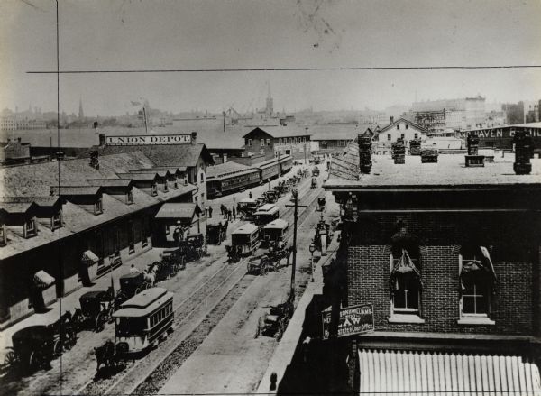 Elevated view of Reed Street from the corner of South Water Street. This depot served the Chicago, Milwaukee and St. Paul, the Wisconsin Central, and the Milwaukee Northern Railways. The depot is on the left, and the downtown is in the far distance. Horse-drawn trolleys and carts are lined up on the curb. Horse-drawn streetcars are coming down the street. There is a sign for a law office on the right.