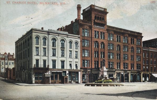 City Hall Square. A statue on a grassy pedestal is in the center of the road in front of the hotel. A grey building is to the left of the hotel on the street corner. Caption reads: "St. Charles Hotel, Milwaukee, Wis."