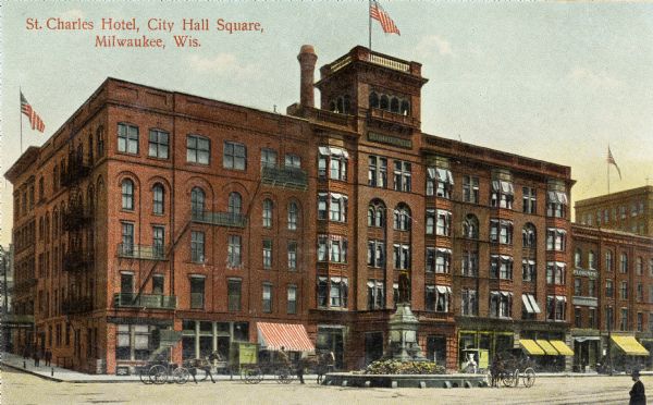 City Hall Square. A statue in a circular area is in the center of the road. Horses and carriages are in the street in front of the hotel. Three flags are on top of the rooftops. Caption reads: "St. Charles Hotel, City Hall Square, Milwaukee, Wis."