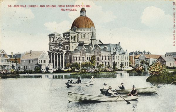 St. Josephat Church and School from Kosciuszko Park. The classic style church with dome is across a body of water. Three boats with boaters are out rowing in the foreground. Along the water's edge are houses and flowering plants. Caption reads: "St. Josephat Church and School from Kosciuszko Park, Milwaukee."