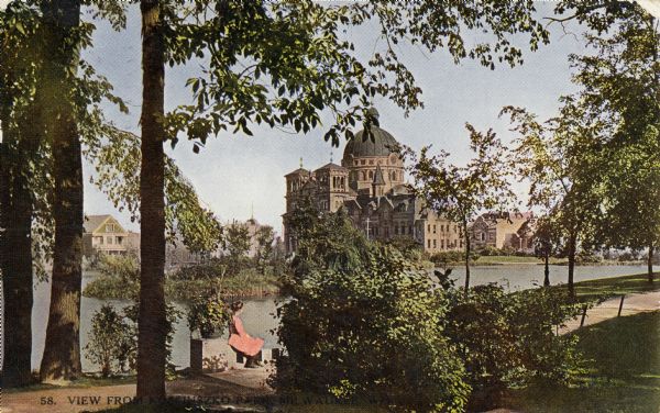 Church and school seen from Kosciuszko Park. Located across a lake, behind some trees and bushes. A woman is sitting on some decorative stones next to the lake. Caption reads: "View from Kosciuszko Park, Milwaukee, Wis."