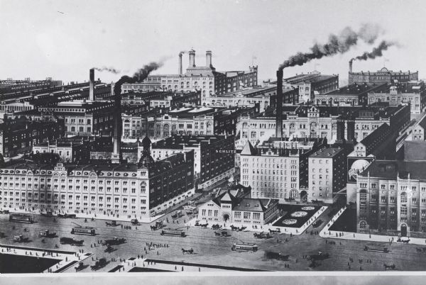Elevated view of brewing company.  A sign for Schlitz is suspended between two of the buildings. Pedestrians, trolleys, and horse-pulled vehicles are on the roads and sidewalks.