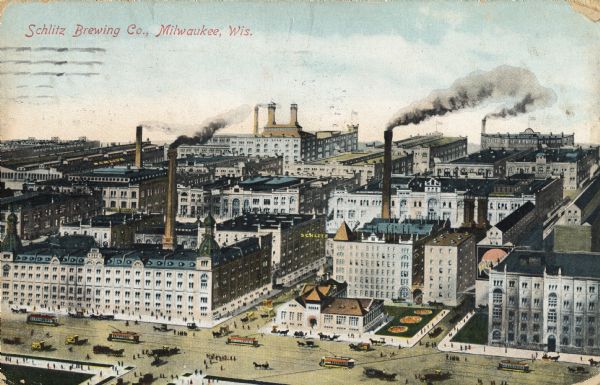 Elevated view of brewing company. A sign for Schlitz is suspended between two of the buildings. Pedestrians, trolleys, and horse-drawn vehicles are on the roads and sidewalks. Caption reads: "Schlitz Brewing Co., Milwaukee, Wis."