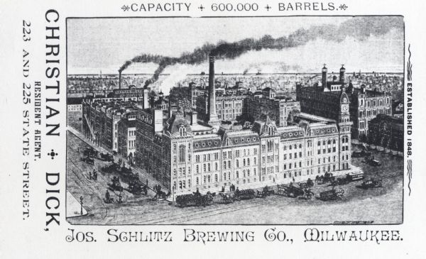 Elevated view of brewing company and surrounding roads. On the road in front of the buildings are horse-drawn carts carrying barrels, a trolley car, and pedestrians. In the distance is Lake Michigan, with ships on the horizon.