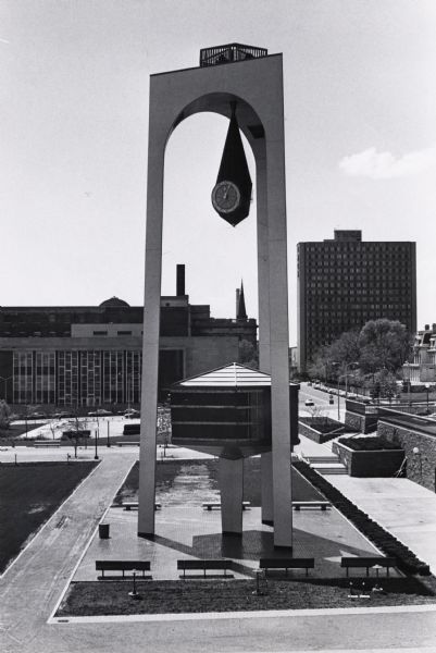 Elevated view of Milwaukee Civic Center Plaza. Entire clock tower is visible, with the plaza around the base, and other buildings in the background.  The clock hangs from the inside roof of the three-legged tower in a teardrop-shaped section. Below the clock is a hexagonal structure on a pedestal, which holds two scenes of historic Milwaukee.  The historic scenes are on stages set in two of the sides of this structure and go into action alternately at regular intervals.