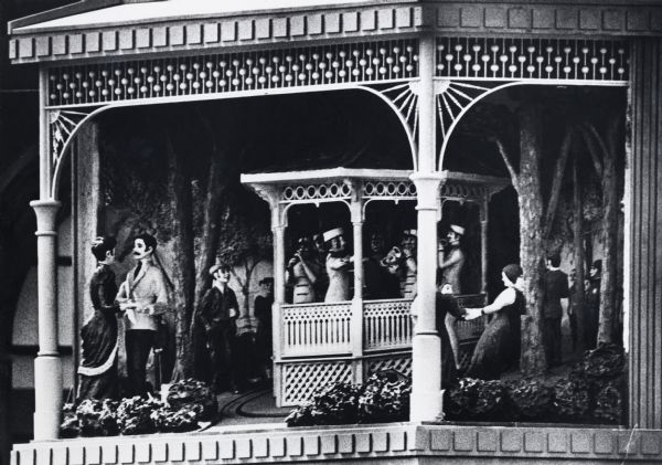 Close-up detail of scene of historic Wisconsin in a hexagonal structure on a pedestal beneath the clock. The scene depicts an outdoor band concert in Milwaukee during the nineteenth century. Five musicians and a conductor perform in a band shell. The scene also includes three girls playing ring-around-the-rosy and a couple waltzing.