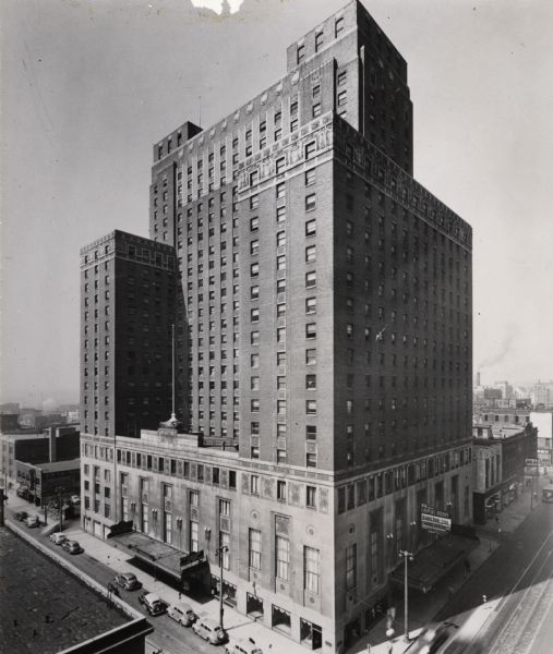 Elevated view of hotel. On the lower right above the entrance is a sign for the Empire Room. Automobiles are parked on the street. A flag pole without a flag is on an upper level of the building on the left above an entrance.