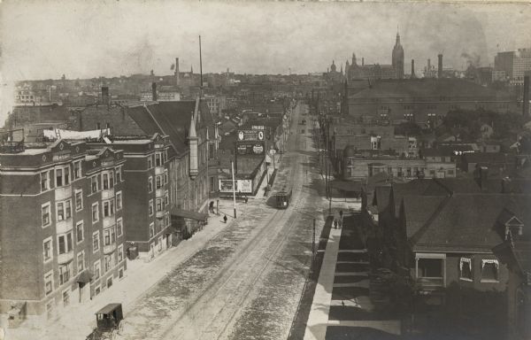 Elevated view looking east along West State Street between Eighth and Ninth Streets.  Numerous buildings line the road, along which cable cars are traveling. A large church building can be seen in the background on the right along the skyline.