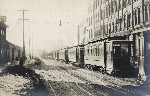 Streetcars are lined up in front of the buildings on the right. On the left people are walking down the sidewalk. A poster hangs from the front of the trolley on the right.