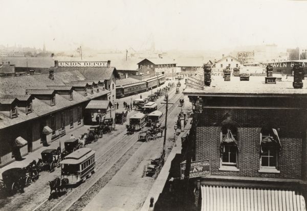 Elevated view of Reed Street from the corner of South Water Street.  This depot served the Chicago, Milwaukee and St. Paul, the Wisconsin Central, and the Milwaukee Northern Railways.  The depot is on the left, and the downtown is in the far distance. Horse-drawn trolleys and carts are lined up on the curb. Horse-drawn streetcars are coming down the street. There is a sign for a law office on the right.