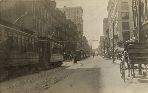Looking down Grand Avenue to West Wisconsin Åvenue. Cable cars are on the left , and a carriage on right near curb. Pedestrians are crossing the street and sidewalk. A large clock is near the intersection.