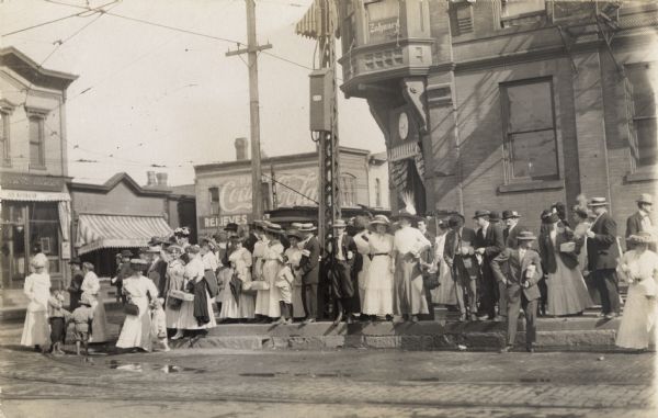 A large crowd of women, children, and men standing on sidewalk and curb. They are probably waiting for the trolley. Many of the people are carrying packages covered in paper and tied up with string.