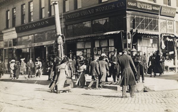 A large group of people are crossing to a street corner. The corner store has a sign saying: "United Cigar Store." The road is paved with cobblestones.