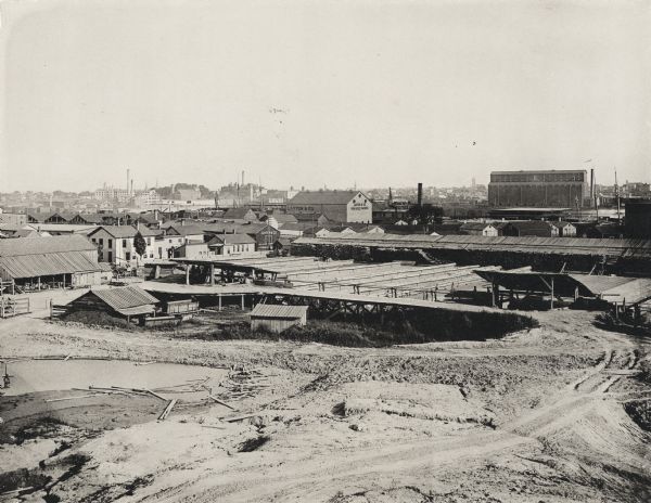 Looking southeast from Clybourn and Fifteenth Streets. An open dirt area is in the foreground, with lumber lying near water on the left, and industrial buildings behind. One of the buildings has a sign that says: Layton & Co. / Pork & Beef Packers." Church spires can be seen in the distance.