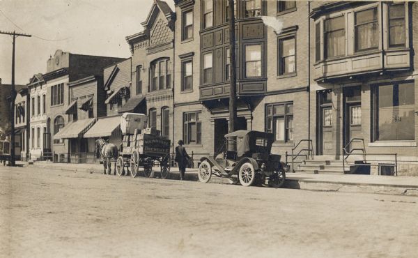 A horse and wagon for the Milwaukee Brewery Co. and a car are parked on the road near the curb.  The road is lined with residences.
