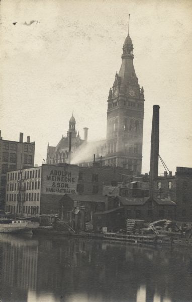 View across river of a boat docked near a building with a sign for Adolph Meinecke & Sons (Manufacturers). In the background is City Hall.