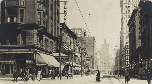 Grand Avenue at 4th Street looking east. The Alhambra Theatre is on the left. Pedestrians are on the streets and sidewalks, and horse-drawn vehicles are on the street. A Gimbels sign is on a rooftop in the background.