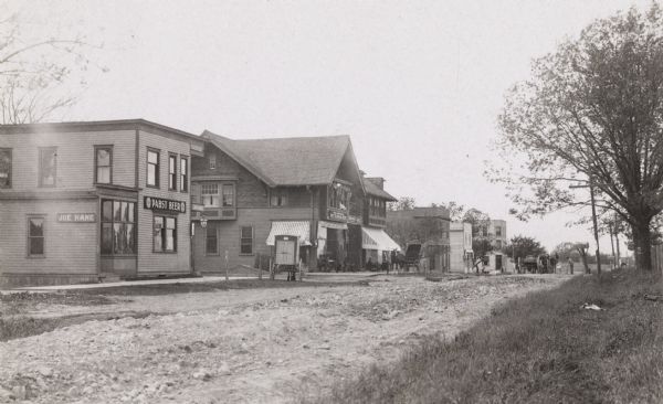 View from dirt road, with buildings grouped on the left, with mailboxes, trees and a fence lining the road on the right. Horse-drawn carts are on the road, and men sitting near the storefronts. The closest building has a sign for Pabst Beer.