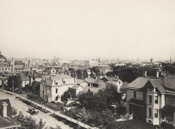 Elevated view southeast from Marquette College.  Homes along street in the foreground with a horse-drawn carriage. Church steeples can be seen in the distance along the skyline.