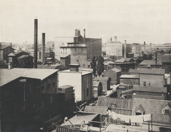 Elevated view looking northwest from Division Street and East Water Street.  Rooftop of building in foreground has laundry hanging from a line. In the background are industrial buildings and chimneys. In the background along a roof line is a sign for "Jos. Schlitz Brewing Co's Bottling Department". Part of a river can be seen near the "Gem Milling Co" building.
