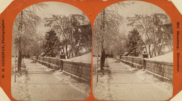 Stereograph.  Winter scene of a residential area, looking down a snowy sidewalk near a retaining wall and a fence in Milwaukee, with a horse-drawn carriage or sleigh in the distance. A flight of steps goes up a hill to the right.