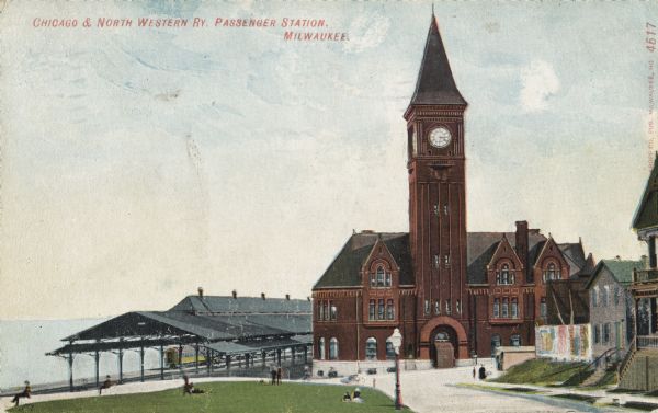 The station with the clock tower is on the right, and the tracks and train shed are on the left. Pedestrians are on the lawn nearby, and houses are on the right of the road that leads to the station. Caption reads: "Chicago & Northwestern Railway Passenger Station, Milwaukee."