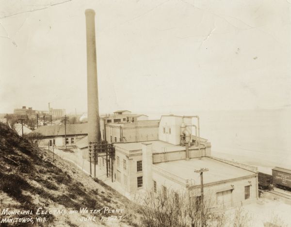 View from a hill looking down on the plant which has several attached buildings, and a large chimney.  To the right are railroad cars along the shoreline of Lake Michigan.