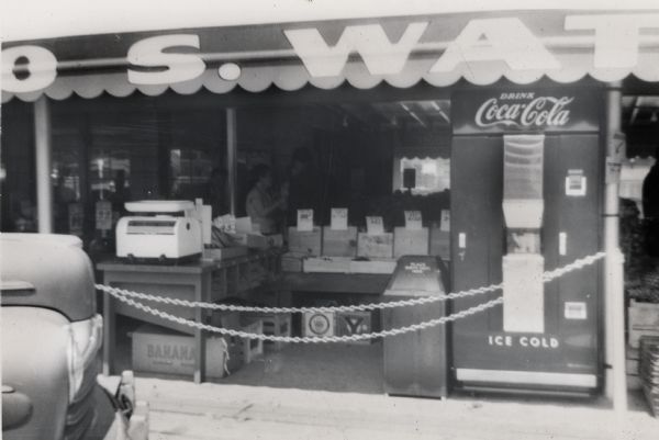 A Coca-Cola vending machine is on the right, near a table with a scale and boxes of fruit, under an awning. On the left is a car near the curb.