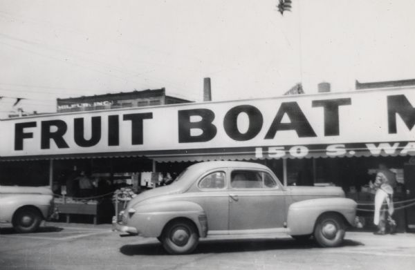 A two-door sedan is parked in front of the market, near a large sign.