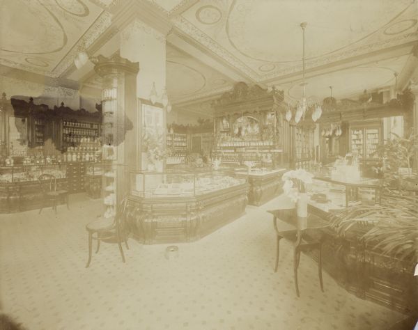 Interior view showing counters, cases, and ornate carved cabinets along the walls display merchandise. Other details include a painted ceiling and column, chairs, carpeting, and potted plants.