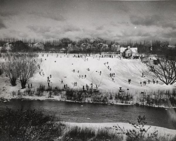 View across river of a large crowd of people on a sledding hill, and also gathered around a shelter building. Houses are in the background atop the hill.