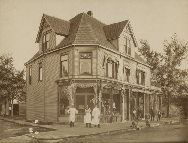 Shops contained on first floor of a Victorian structure, one of which belongs to a woman named Mary. Standing in front are her husband and two sons. Further down the sidewalk in front of another storefront is a man and three boys.  Three children sit at the curb.  The wheels of a cart are on the far right.