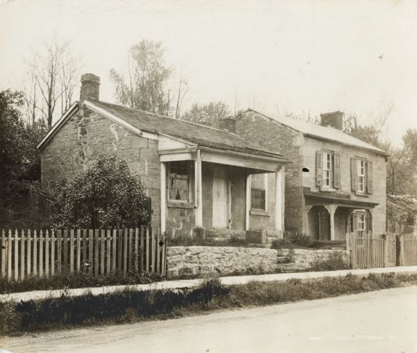 On Shakerag Street. Two houses, both of stone, behind a wood fence and a low stone wall along a sidewalk.  The house on the right is two stories, the left is a one story structure.