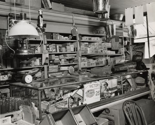 Interior view of a counter and shelves displaying merchandise.  Boxes of various items line the walls, pails hang from the ceiling, and a lamp hangs on the left.