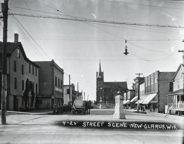 A church is in the distance at the end of the road.  On either side of the street are homes and stores, with a street light suspended above the street in the foreground.  An automobile and a horse-drawn cart with a large barrel are on the left, and pedestrians are on the sidewalk  on the right.  One person has a bicycle.  In the middle of the road is a traffic post with advertisements.  Before the church is a statue commemorating the first Swiss colony in 1845.