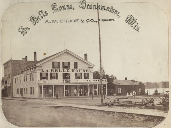 A summer resort, situated on the banks of Fowler and La Belle Lakes, in the village of Oconomowoc, thirty miles from Milwaukee on the St. Paul R.R. Men stand on the porch, with people on the balcony above them. A lake can be seen in the background.
