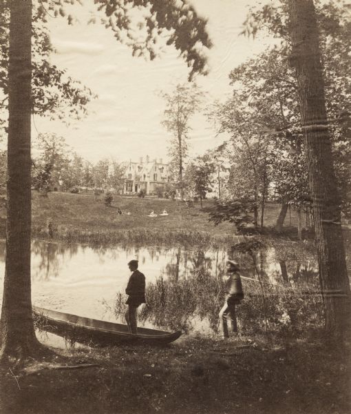 Two men are in the foreground on a shoreline, one standing in a boat.  In the background on the other shoreline are adults and children sitting in the grass. There is a large, ornately decorated white house behind them.