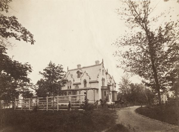 A large, white brick building is in the middle center, surrounded by trees.  A smaller white building is in the right background.  In front of the building is a man driving two horses and a cart. A man is standing along a fence, and in the background is a lake.