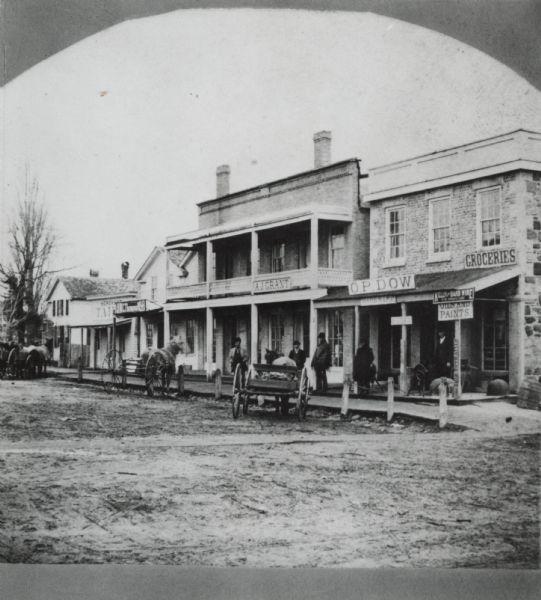 Storefronts and homes along dirt street. Three horse-drawn vehicles are tethered to posts in front of a board sidewalk. Men stand on the sidewalk in front of two stores which have a balcony.