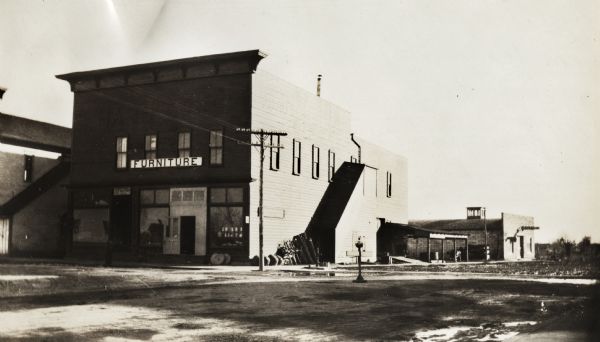 Two-story building from across a dirt road, with a small building behind it on the right. Some goods are stacked along the exterior wall near an exterior covered stairway. A large "Furniture" sign is above the entrance.