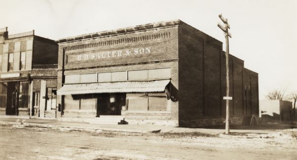 View across road of two-story brick building with a small awning over the front.  Located on the corner of a dirt road.  The awning has "General Merchandise" written on the top, and the letters placed on the second floor read "R.B. Salter & Son."